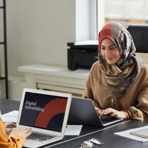 Two women in hijabs work on laptops in a bright office. Perfect for business and diversity themes.